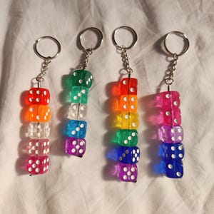 May include: Four keychains each featuring a vertical stack of five translucent, colorful dice with white pips. The dice are connected by a metal pin and attached to a silver-toned metal split ring by a short chain. The colors include red, orange, clear, pink, green, blue, purple, and yellow.