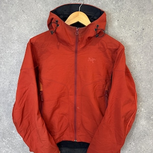 May include: A rust-orange hooded jacket featuring a full-length front zipper, a high collar, and adjustable drawstrings on the hood. The garment includes two side pockets with vertical zippers and a small embroidered logo on the upper chest area.