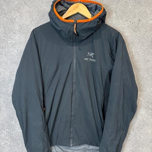 May include: A dark charcoal gray hooded jacket featuring a full-length front zipper, long sleeves, and a bright orange trim along the edge of the hood. The Arc'teryx logo is printed in white on the upper left chest area of the garment.
