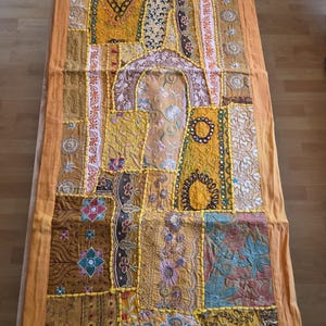 Puede incluir: Un camino de mesa rectangular con un diseño de patchwork de varios paneles de tela en tonos amarillo, naranja y marrón. El textil incluye bordados intrincados, hilos metálicos, pequeños espejos circulares y patrones florales en la superficie.