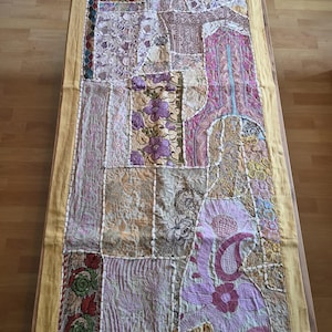 May include: A rectangular table runner featuring a patchwork design of various embroidered fabrics in shades of purple, pink, gold, and cream. The runner is bordered by a solid yellow fabric and displays intricate floral and paisley patterns.