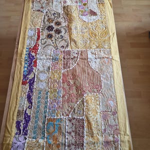 May include: A rectangular table runner featuring a patchwork design of various embroidered fabric panels in shades of gold, yellow, cream, and multi-colored floral patterns. The piece is bordered by a solid yellow fabric edge and white stitching.