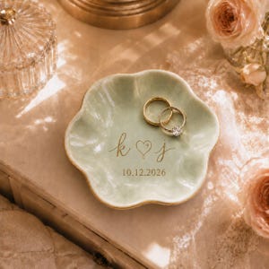 May include: A pale green, flower-shaped ceramic trinket dish featuring gold-colored cursive lettering that reads k heart j and the date 10.12.2026. Two gold-toned metal rings, one with a clear stone, rest inside the dish on the smooth surface.