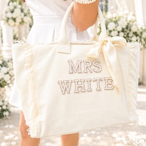 May include: A cream-colored canvas tote bag featuring frayed edges and a satin ribbon tied to the handle. The front displays the text MRS WHITE in block letters embellished with small white pearls, creating a textured and decorative appearance.