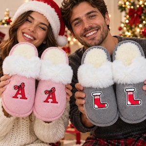May include: A pair of light pink slippers featuring a red letter A with a polka dot bow and a pair of gray slippers featuring a red and white Christmas stocking. Each slipper has a white faux fur trim around the opening and a soft interior lining.