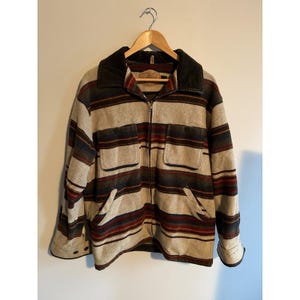 May include: A zip-up jacket featuring a horizontal striped pattern in shades of beige, dark red, and dark green. The garment includes a dark brown collar, two chest pockets with flaps, two lower hand pockets, and long sleeves with buttoned cuffs.