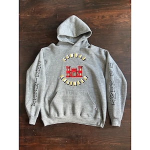 May include: A heather grey hooded sweatshirt featuring a red castle emblem and the text "COMBAT ENGINEER" arched above and below it. The left sleeve displays the word "ENGINEER" and the right sleeve displays "US ARMY" in black lettering.