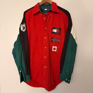 May include: A long-sleeve button-down shirt featuring a red center panel, black and green sleeves, and white accents. It displays a Tommy Hilfiger logo, a Norwegian flag, a Canadian flag, and a patch of a skier on the right sleeve and collar.