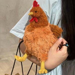 以下が含まれることがあります： 質感のある茶色の毛並み、鮮やかな赤色のトサカと肉垂、黄色のくちばし、黄色の布製の足を持つ鶏のぬいぐるみ。このぬいぐるみは丸みを帯びた体型で、小さな黒い目をしており、全体が柔らかく毛足の長い素材で作られています。