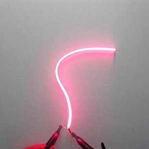 Puede incluir: Una tira de luz de neón rosa flexible y brillante con una curva suave. La tira está conectada en su base a dos conectores eléctricos de color oscuro, que suministran energía al tubo iluminado, creando una luz rosa brillante y continua.