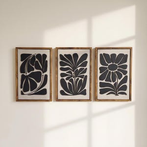 May include: A set of three rectangular wall art pieces featuring black organic floral shapes mounted on a light beige fabric background, each enclosed within a medium-toned wooden frame with a visible grain, arranged horizontally in a row.