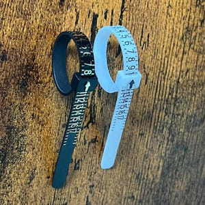 Könnte beinhalten: Ein schwarzer Ringgrößenmesser und ein weißer Ringgrößenmesser, beide mit einem Schnallenmechanismus mit Pfeilmarkierung und aufgedruckten numerischen Skalen von 1 bis 17, zur Messung des Fingerumfangs zur Bestimmung der Ringgröße.