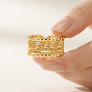 Op de afbeelding: Een goudkleurige ring met meerdere banden en een breed ontwerp dat bestaat uit verschillende gestapelde horizontale banden. De buitenste banden hebben een kralentextuur, terwijl de binnenste banden bestaan uit gladde en fijn getextureerde draden.