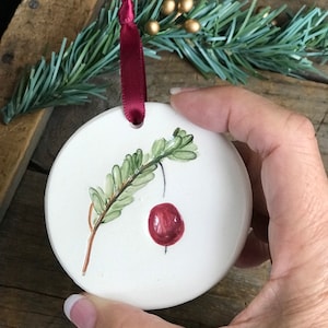 May include: A round, white ceramic Christmas ornament with a hand-painted cranberry and branch design. The ornament has a burgundy ribbon for hanging. Green pine needles and gold berries are in the background.
