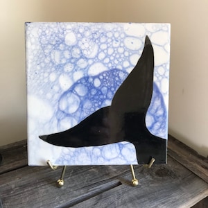 May include: A square ceramic tile with a blue and white bubble design. A black silhouette of a whale tail is painted on the tile.
