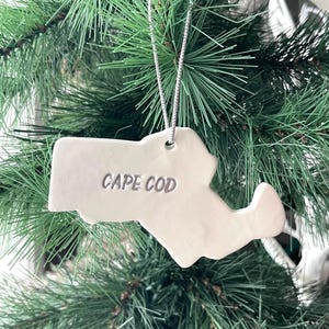 May include: A white ceramic ornament shaped like the map of Cape Cod, featuring the text CAPE COD embossed in the center. The ornament is suspended by a thin, silver-colored cord, designed for hanging on a tree or other decorative display.