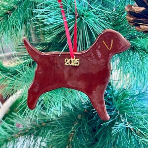 Pu&ograve; includere: Una decorazione natalizia in ceramica a forma di cane, di colore rosso scuro lucido con una finitura liscia. Un ciondolo in metallo dorato &egrave; appeso a un nastro rosso e riporta l'anno 2025. &Egrave; visibile un semplice contorno dorato dell'orecchio.