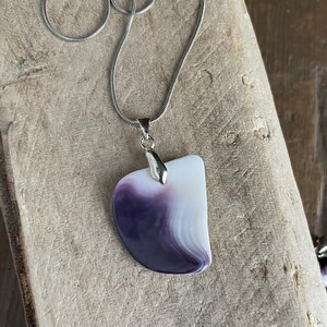 May include: A silver necklace with a pendant featuring a white and purple stone. The pendant has a curved, asymmetrical shape with a polished finish. The chain is a thin, silver snake chain.