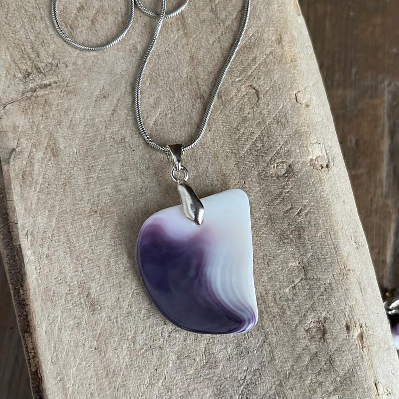 May include: A silver necklace with a pendant featuring a white and purple stone. The pendant has a curved, asymmetrical shape with a polished finish. The chain is a thin, silver snake chain.