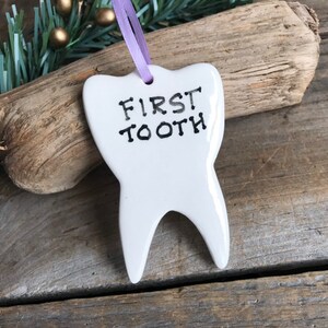 May include: A white ceramic tooth-shaped ornament with the text "FIRST TOOTH" printed in black ink. The ornament features a smooth, glossy finish and is suspended by a thin, light purple ribbon attached through a hole at the top center.