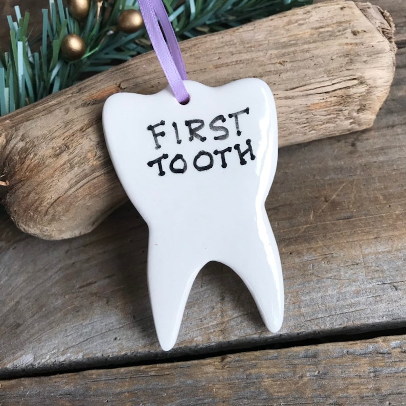 May include: A white ceramic tooth-shaped ornament with the text "FIRST TOOTH" printed in black ink. The ornament features a smooth, glossy finish and is suspended by a thin, light purple ribbon attached through a hole at the top center.