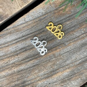 May include: Two small metal charms shaped like the number 2026, one in a gold-tone finish and one in a silver-tone finish. Each charm features a small circular loop at the top for attachment, with the digits clearly embossed in a bold font.
