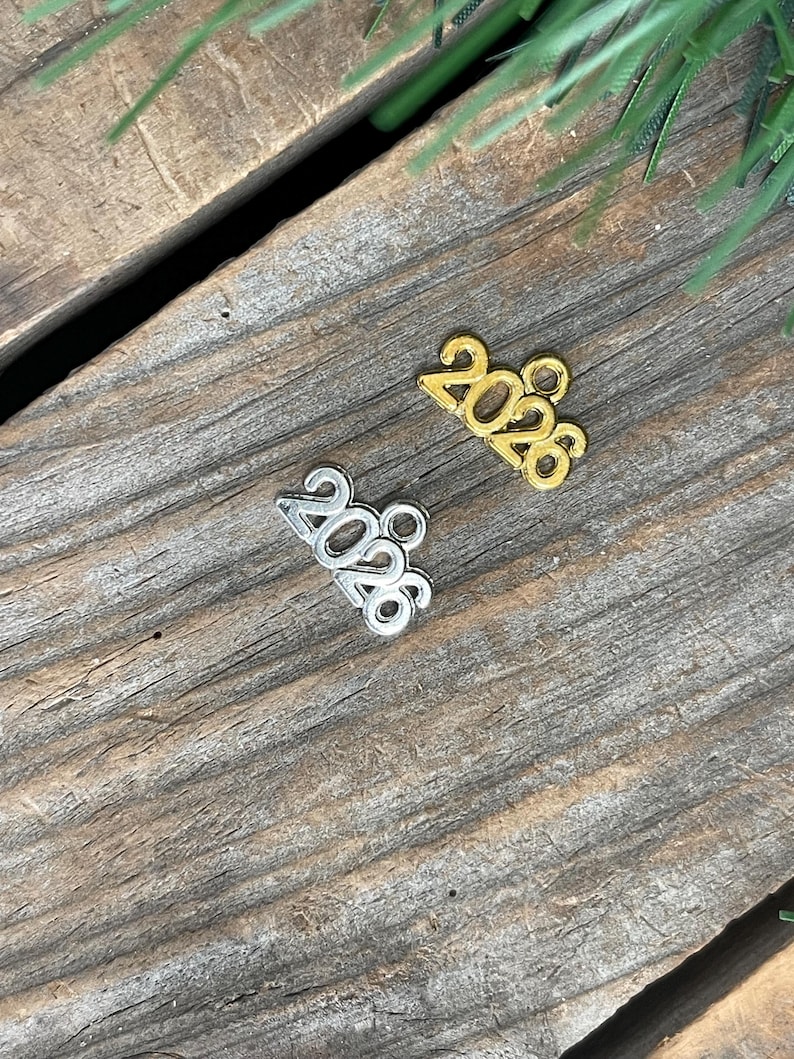 May include: Two small metal charms shaped like the number 2026, one in a gold-tone finish and one in a silver-tone finish. Each charm features a small circular loop at the top for attachment, with the digits clearly embossed in a bold font.