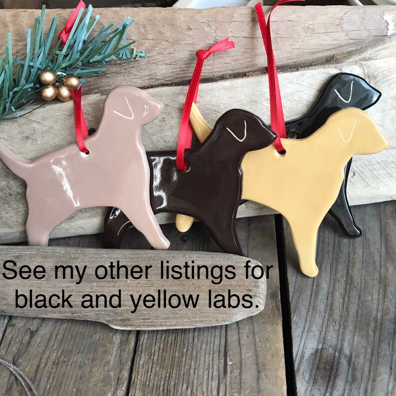 Pu&ograve; includere: Quattro ornamenti a forma di cane in marrone chiaro, marrone scuro, giallo e nero, ciascuno con un nastro rosso per appenderli. Il testo "See my other listings for black and yellow labs." &egrave; stampato sulla superficie in legno sotto gli ornamenti.