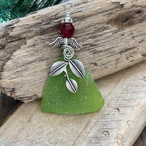 Pode incluir: Enfeite de anjo artesanal com um corpo de vidro do mar verde. O anjo tem asas prateadas e um design de folhas, encimado por uma conta facetada vermelha. O enfeite &eacute; suspenso por um la&ccedil;o de arame prateado, perfeito para decora&ccedil;&atilde;o festiva.