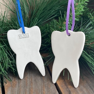 May include: Two white ceramic tooth-shaped ornaments. One ornament features a blue ribbon and a silver charm with the text 2023. The other ornament features a purple ribbon. Both items are smooth, glossy, and shaped like a human molar.