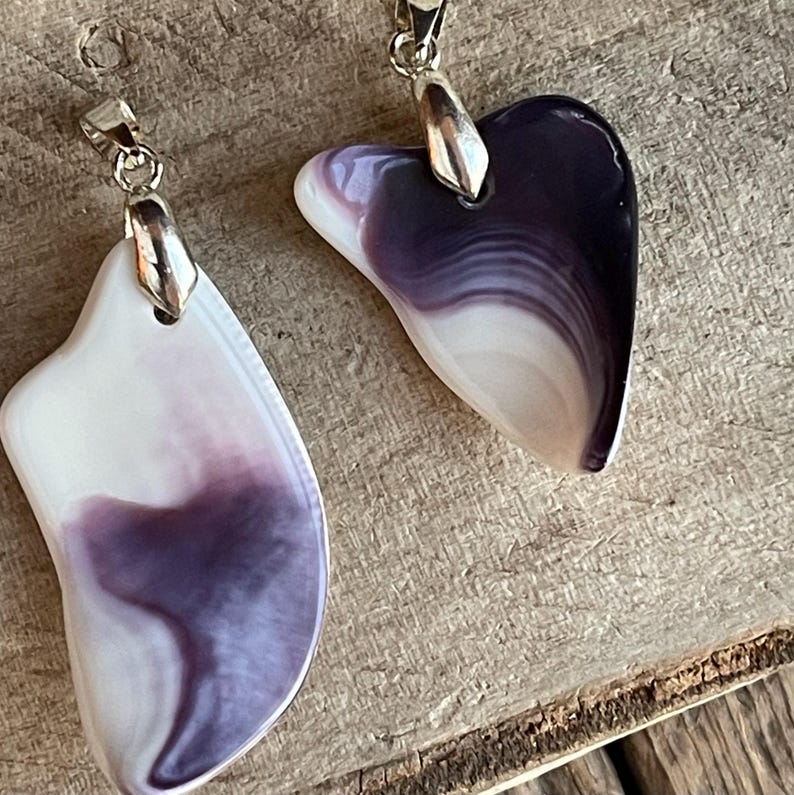 May include: Two pendants with silver-colored bails. One is a heart shape, the other is an irregular shape. Both are white with purple marbling. The pendants are displayed on a wooden surface.