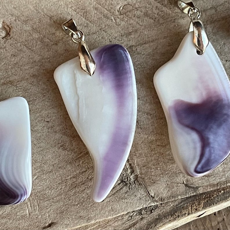 May include: Close-up of several pendants with a white and purple color scheme. Each pendant has a unique shape and a silver-colored bail. The pendants are displayed on a wooden surface.