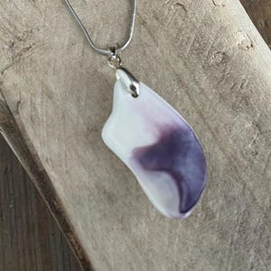 May include: A pendant necklace featuring a white and purple abstract design. The pendant is attached to a silver chain. The necklace is displayed on a wooden surface.
