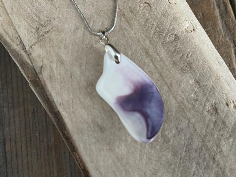 May include: A pendant necklace featuring a white and purple abstract design. The pendant is attached to a silver chain. The necklace is displayed on a wooden surface.