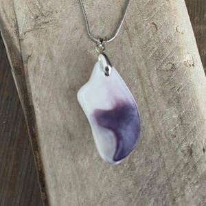 May include: A pendant necklace featuring a white and purple abstract design. The pendant is attached to a silver-colored chain. The pendant has a smooth, polished surface and a unique, organic shape. The necklace is displayed on a wooden surface.