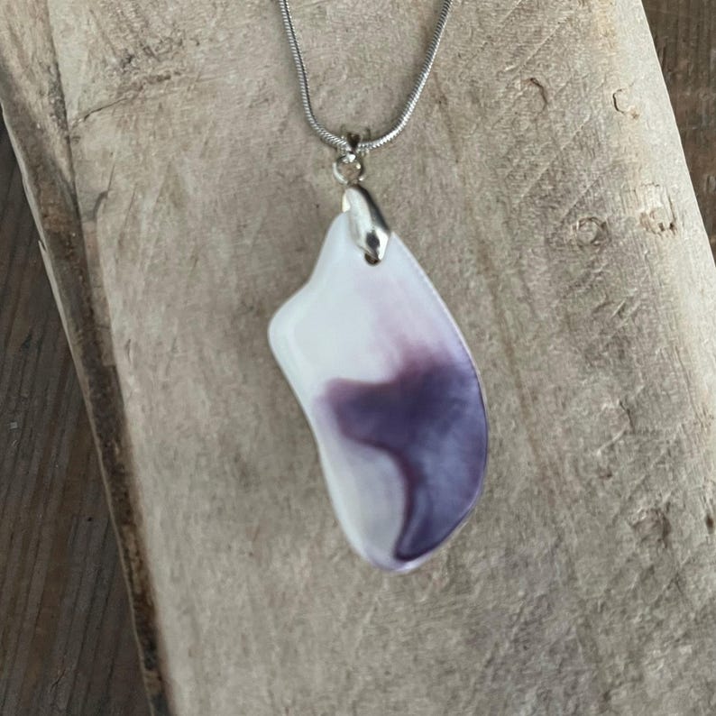 May include: A pendant necklace featuring a white and purple abstract design. The pendant is attached to a silver-colored chain. The pendant has a smooth, polished surface and a unique, organic shape. The necklace is displayed on a wooden surface.