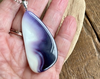 Pode incluir: Um pingente em forma de lágrima com uma superfície lisa e polida, apresentando um gradiente de cores violeta profundo, lavanda e branco creme. O pingente está preso a um suporte de metal prateado no topo, exibindo um padrão espiralado.