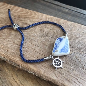 May include: A blue and white porcelain charm with a silver ship's wheel charm hangs from a navy blue cord bracelet.
