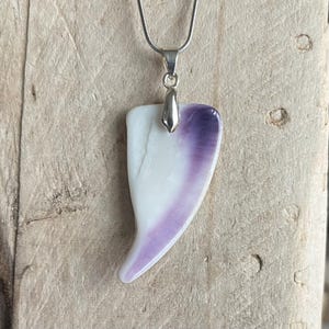 May include: A pendant necklace featuring a white and purple shell-shaped pendant. The pendant is suspended from a silver-colored chain. The pendant has a smooth, polished surface and a unique, organic shape.