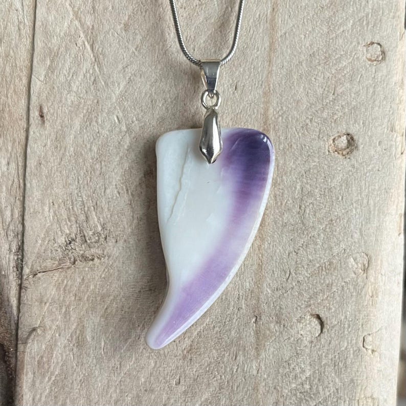 May include: A pendant necklace featuring a white and purple shell-shaped pendant. The pendant is suspended from a silver-colored chain. The pendant has a smooth, polished surface and a unique, organic shape.
