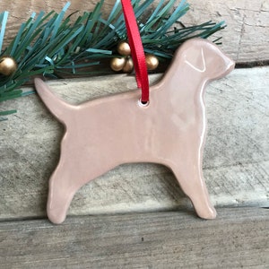 Pu&ograve; includere: Un ornamento in ceramica color beige chiaro lucido a forma di cane di profilo. L'ornamento presenta una superficie liscia e riflettente e un piccolo foro nella parte superiore, attraverso il quale &egrave; infilato un sottile nastro rosso vivo.