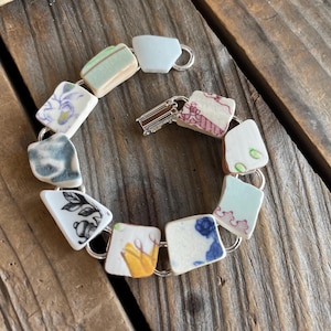 May include: A bracelet composed of ten irregular ceramic shards connected by silver-toned metal jump rings. The pieces feature various patterns including floral motifs, abstract shapes, and solid colors like white, blue, green, yellow, and pink.