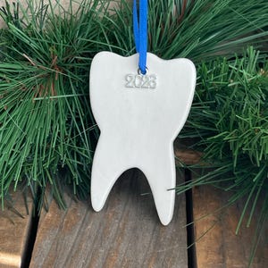 May include: A white ceramic tooth-shaped ornament featuring a silver-toned metal charm engraved with the year 2026. The ornament is suspended by a bright blue ribbon, creating a distinct contrast against the smooth, glossy white surface.