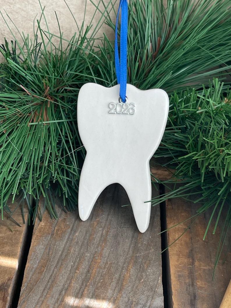 May include: A white ceramic tooth-shaped ornament featuring a silver-toned metal charm engraved with the year 2026. The ornament is suspended by a bright blue ribbon, creating a distinct contrast against the smooth, glossy white surface.
