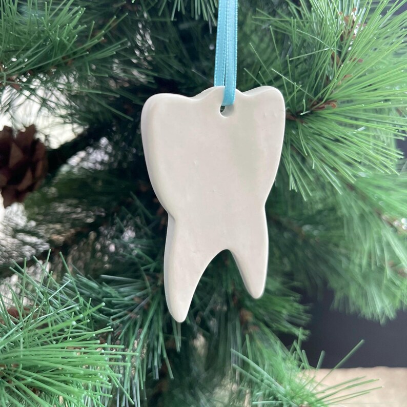 May include: A white ceramic tooth-shaped ornament with a smooth, matte finish. The ornament features a small hole at the top, through which a light blue satin ribbon is threaded for hanging. The shape mimics a human molar with two roots.
