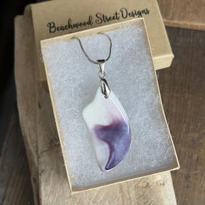 May include: A pendant necklace with a white and purple abstract design. The pendant is on a silver chain and is displayed in a small gift box. The box has the text "Beachwood Street Designs" printed on it.
