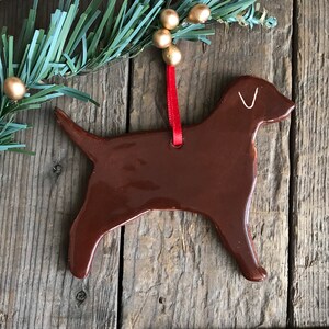 Pu&ograve; includere: Un ornamento in ceramica marrone lucido a forma di cane Labrador. L'ornamento presenta un semplice contorno bianco di un orecchio sulla testa ed &egrave; appeso a un sottile nastro rosso. La superficie &egrave; liscia e riflettente.