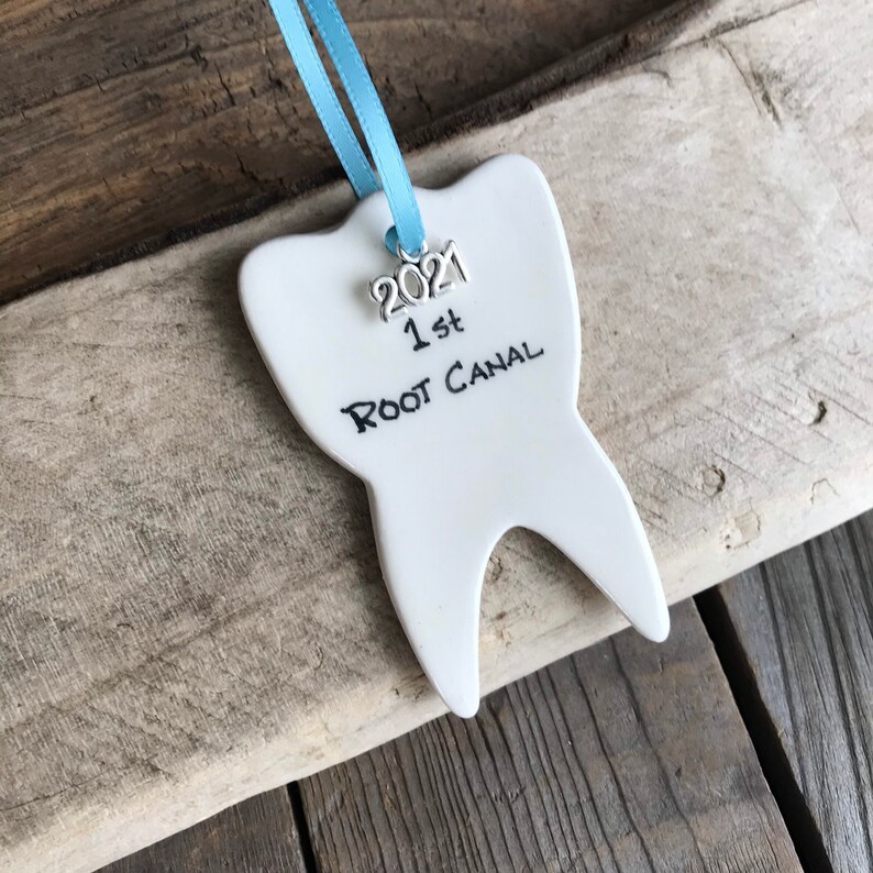 May include: A white ceramic tooth-shaped ornament hanging from a light blue ribbon. The ornament features a silver-toned metal charm with the year 2021 and the black handwritten text 1st ROOT CANAL printed on the front surface of the tooth.
