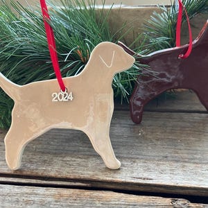 Pu&ograve; includere: Due ornamenti in ceramica a forma di cane con finitura lucida. Uno &egrave; beige chiaro e l'altro &egrave; marrone scuro. Ogni ornamento &egrave; appeso a un nastro rosso e presenta un piccolo ciondolo argentato a forma di numero 2024 sul passante.