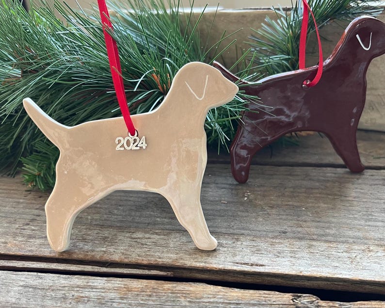 Pu&ograve; includere: Due ornamenti in ceramica a forma di cane con finitura lucida. Uno &egrave; beige chiaro e l'altro &egrave; marrone scuro. Ogni ornamento &egrave; appeso a un nastro rosso e presenta un piccolo ciondolo argentato a forma di numero 2024 sul passante.